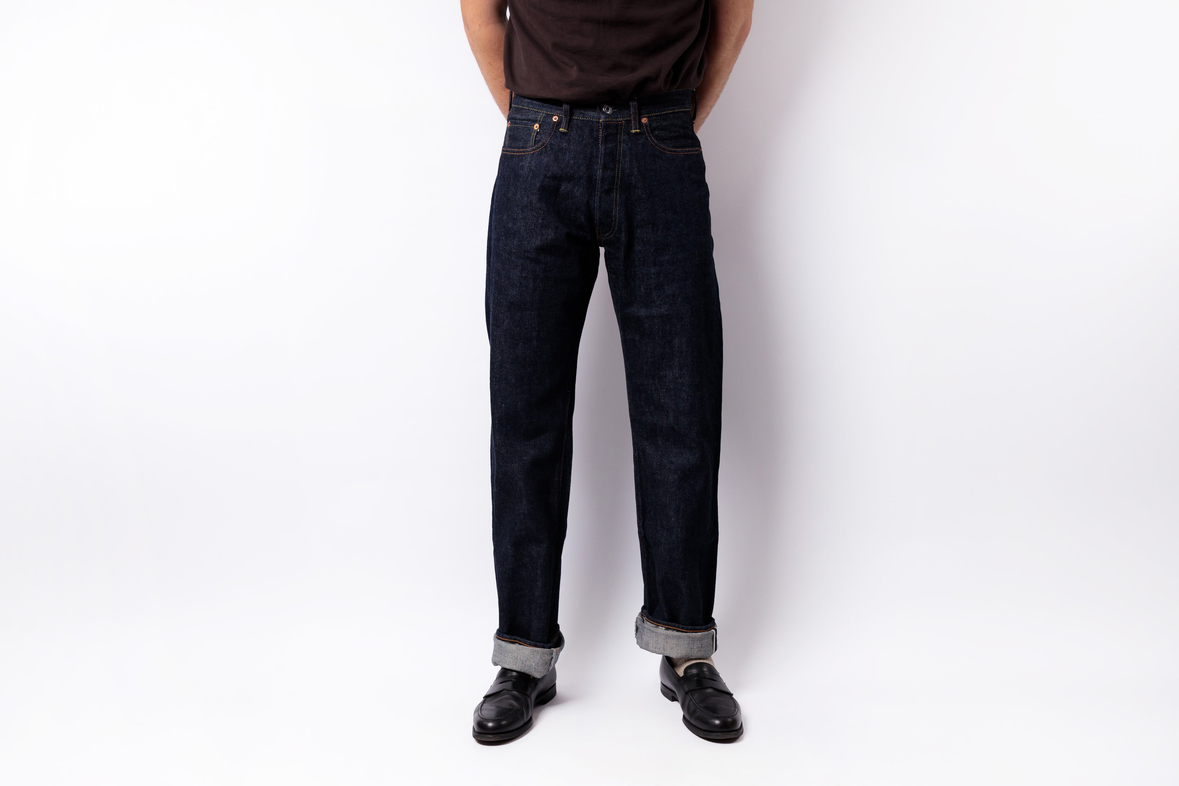 A person wearing dark indigo denim jeans with rolled cuffs, paired with a dark brown t-shirt and black leather shoes, standing against a plain white studio background.