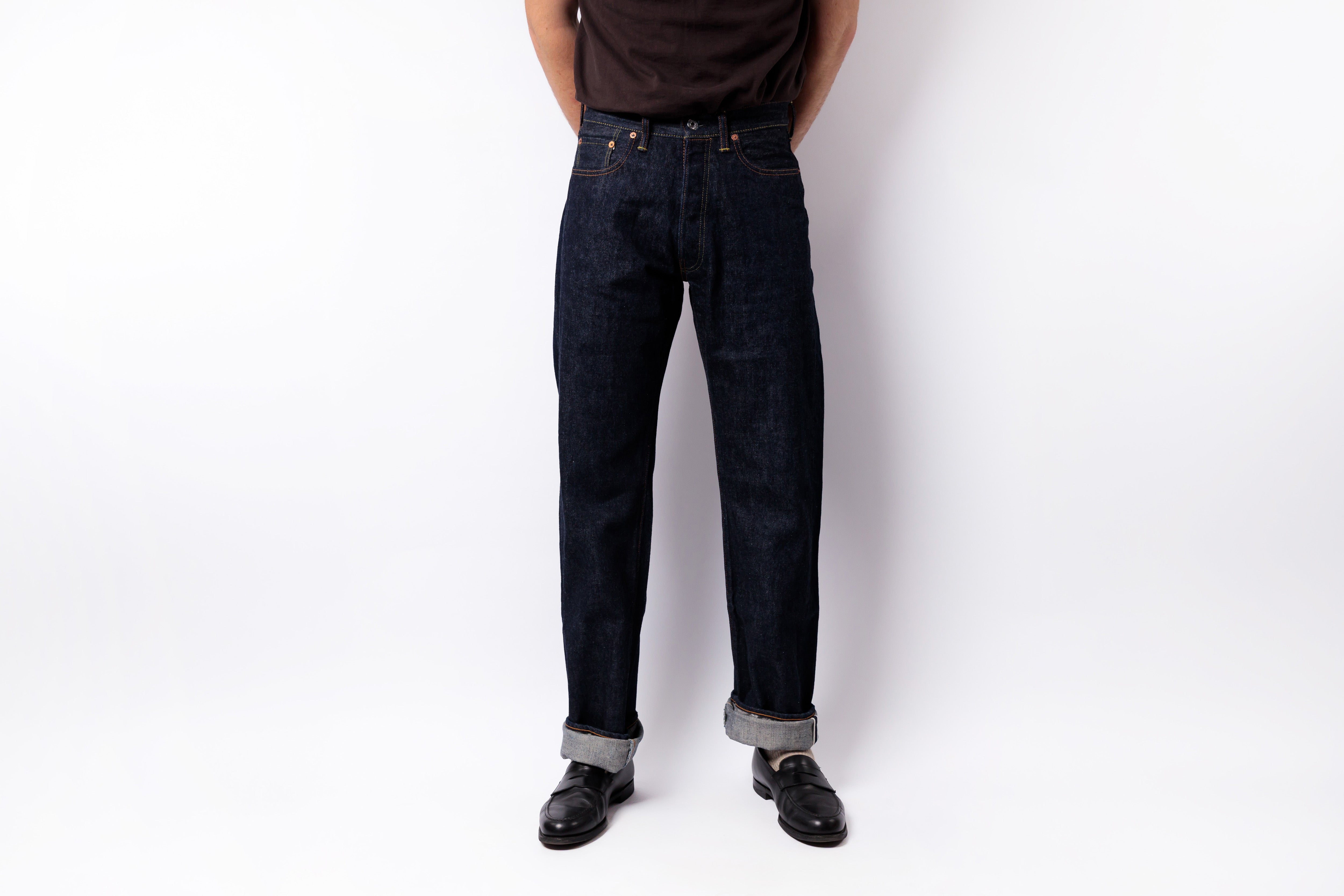 A person wearing dark indigo denim jeans with rolled cuffs, paired with a dark brown t-shirt and black leather shoes, standing against a plain white studio background.