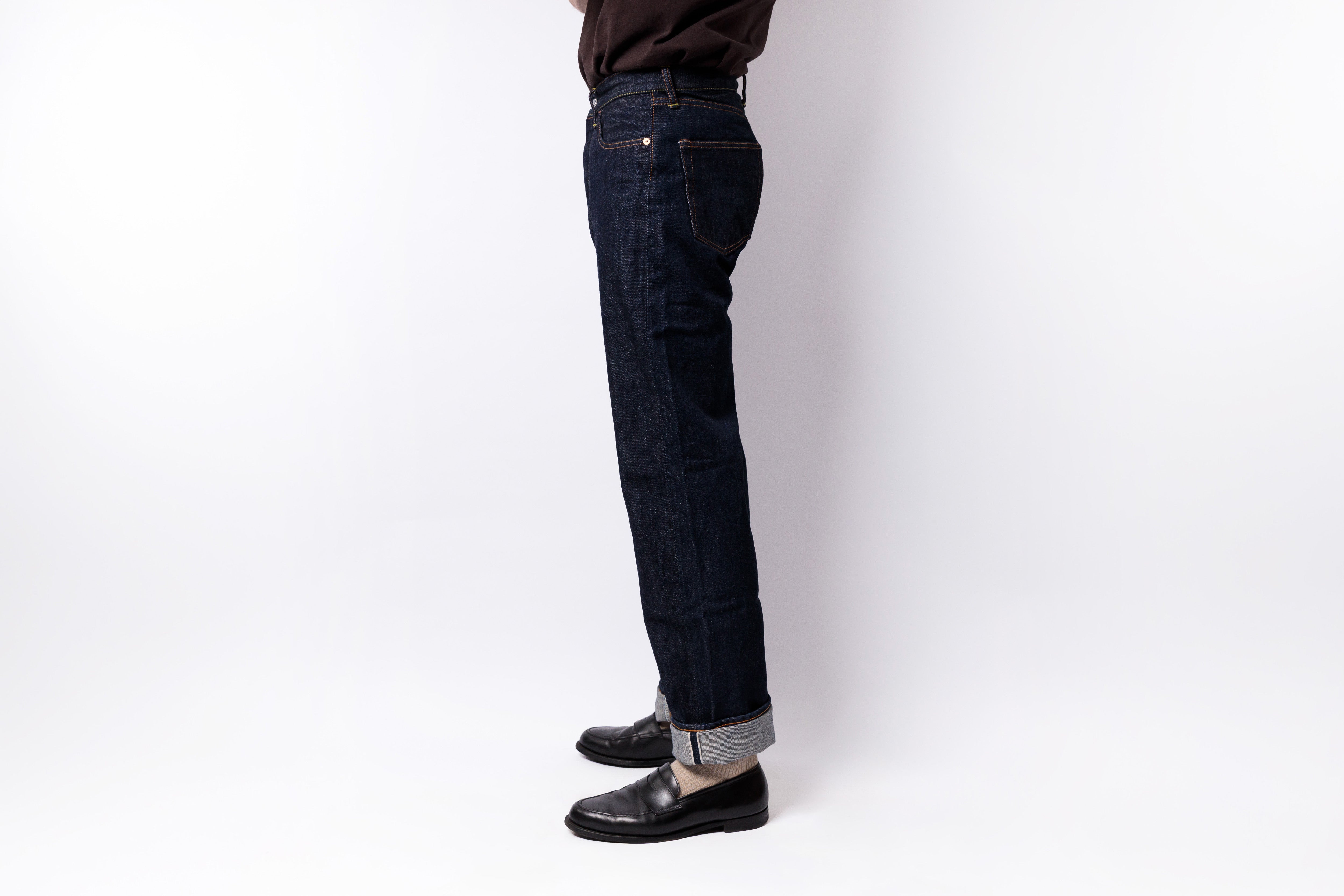 A side profile view of a person wearing dark indigo denim jeans with rolled cuffs, paired with black leather loafers and a dark brown shirt, standing against a plain white studio background.