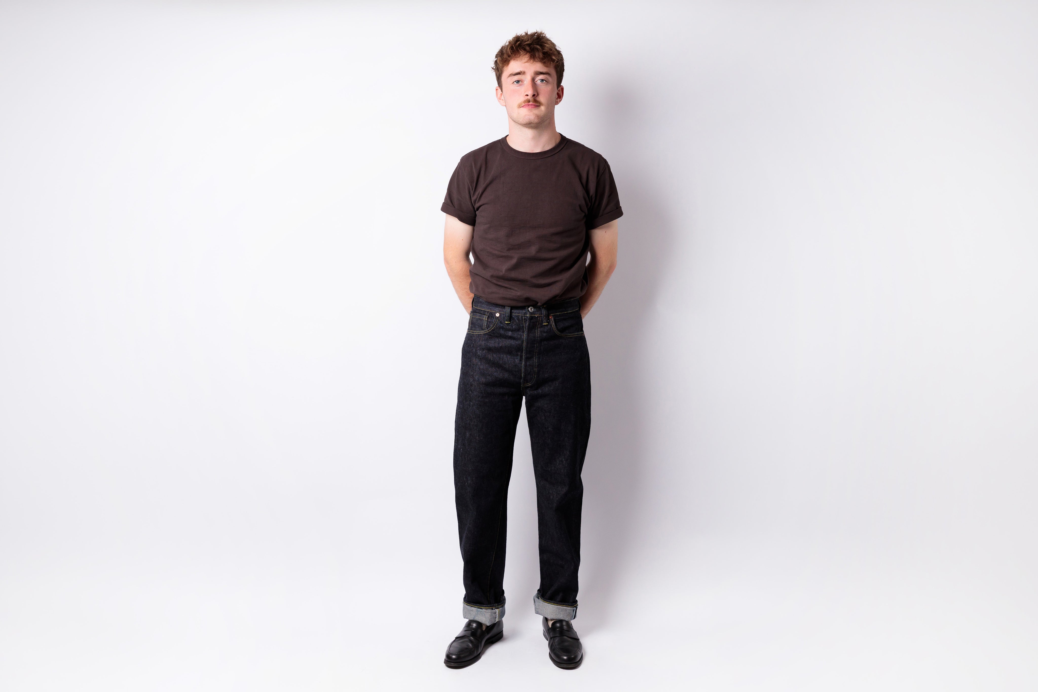 A man standing against a light gray studio background, wearing a dark brown crew-neck t-shirt, dark wash jeans, and black shoes.