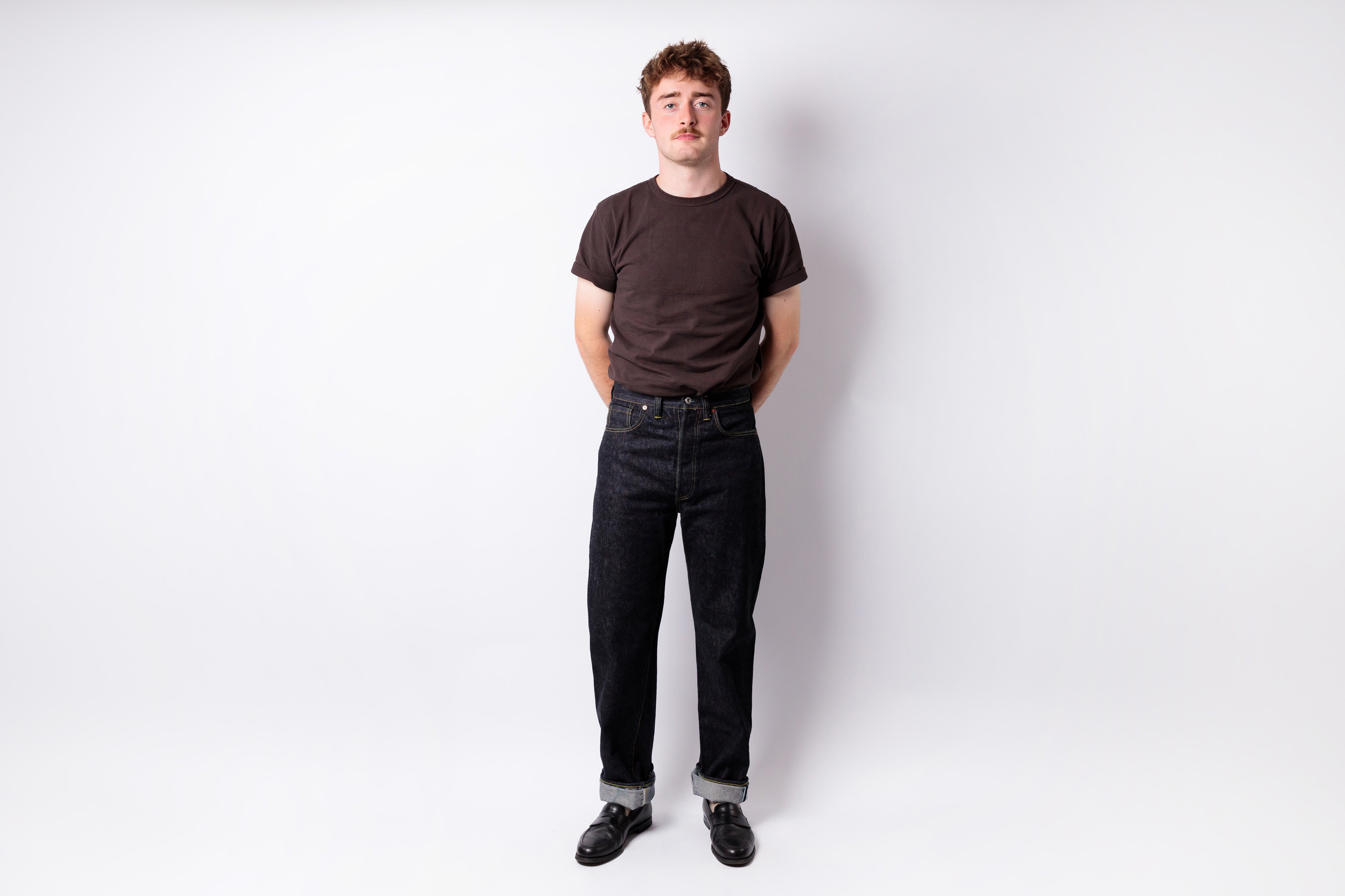 A man standing against a light gray studio background, wearing a dark brown crew-neck t-shirt, dark wash jeans, and black shoes.