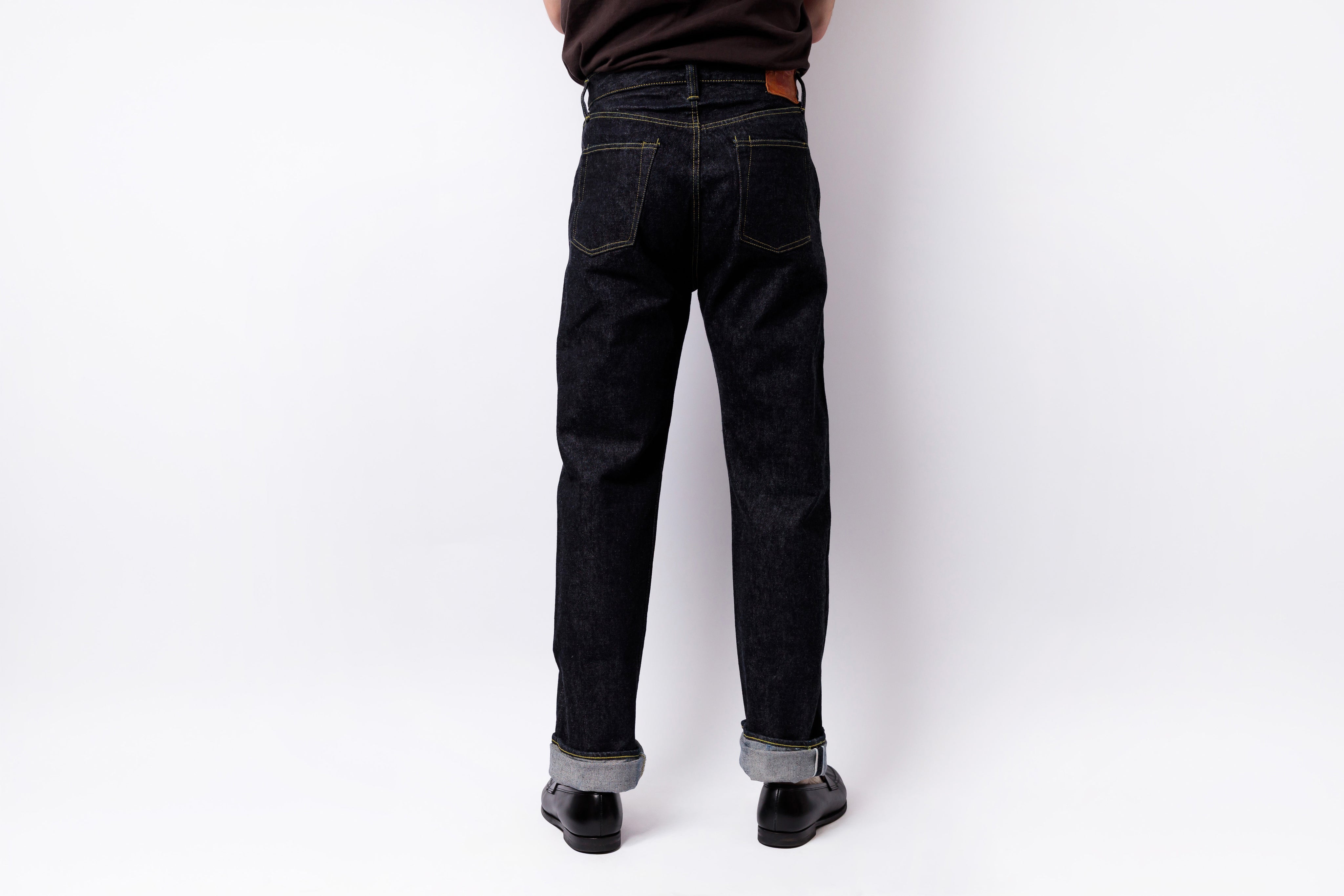 Rear view of a person wearing dark indigo denim jeans with rolled cuffs, black shoes, and a dark brown shirt against a plain white studio background.