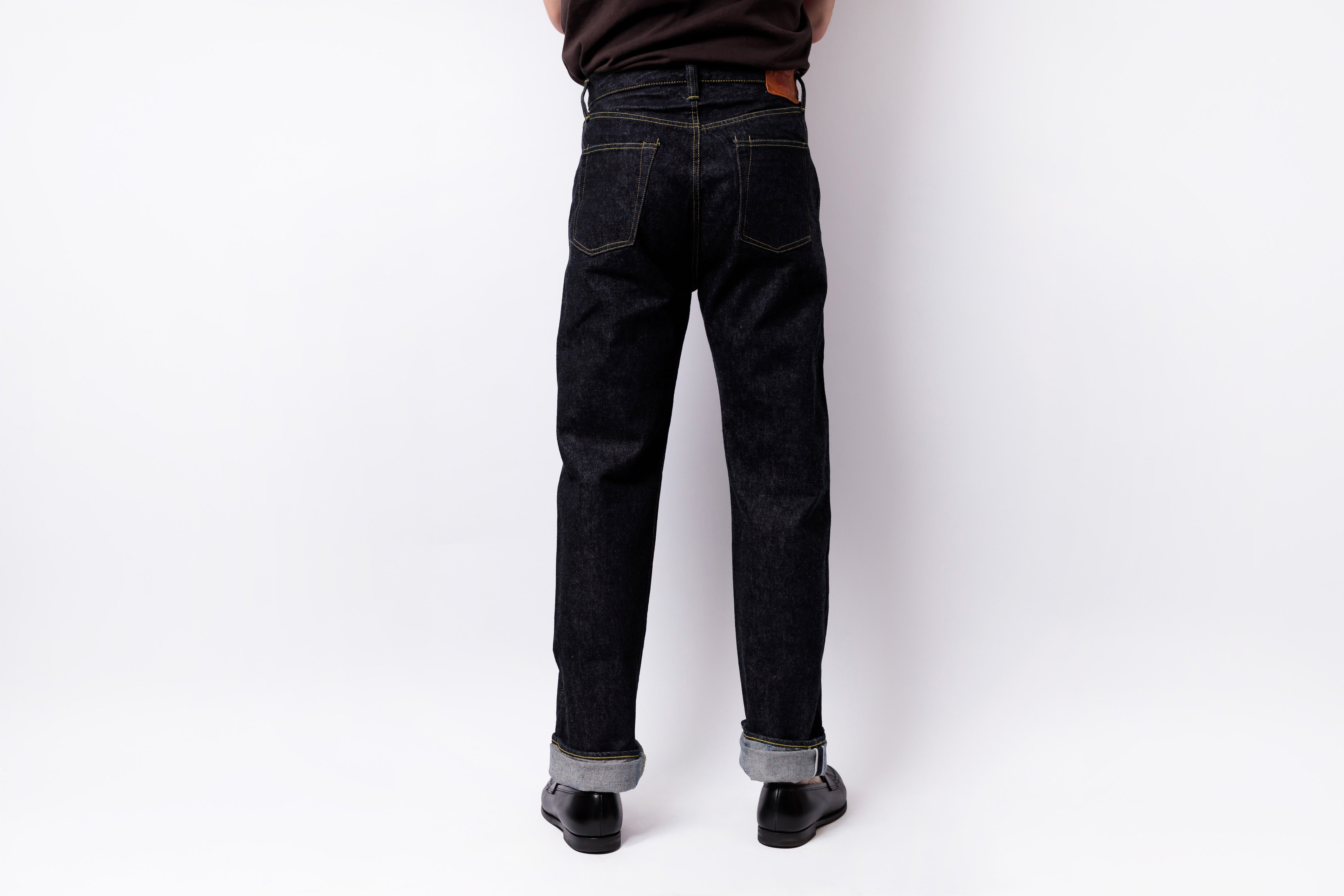 Rear view of a person wearing dark indigo denim jeans with rolled cuffs, black shoes, and a dark brown shirt against a plain white studio background.