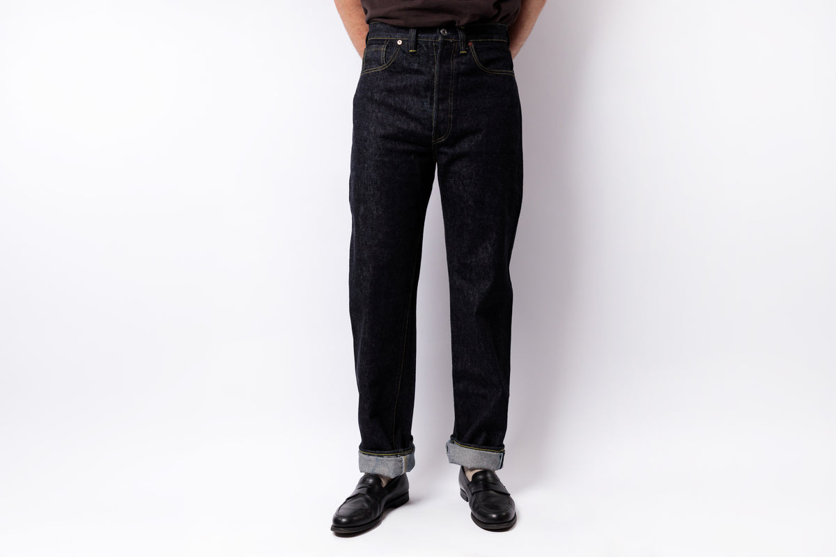 A person wearing dark indigo denim jeans with rolled cuffs, paired with black leather shoes, standing against a plain white studio background.