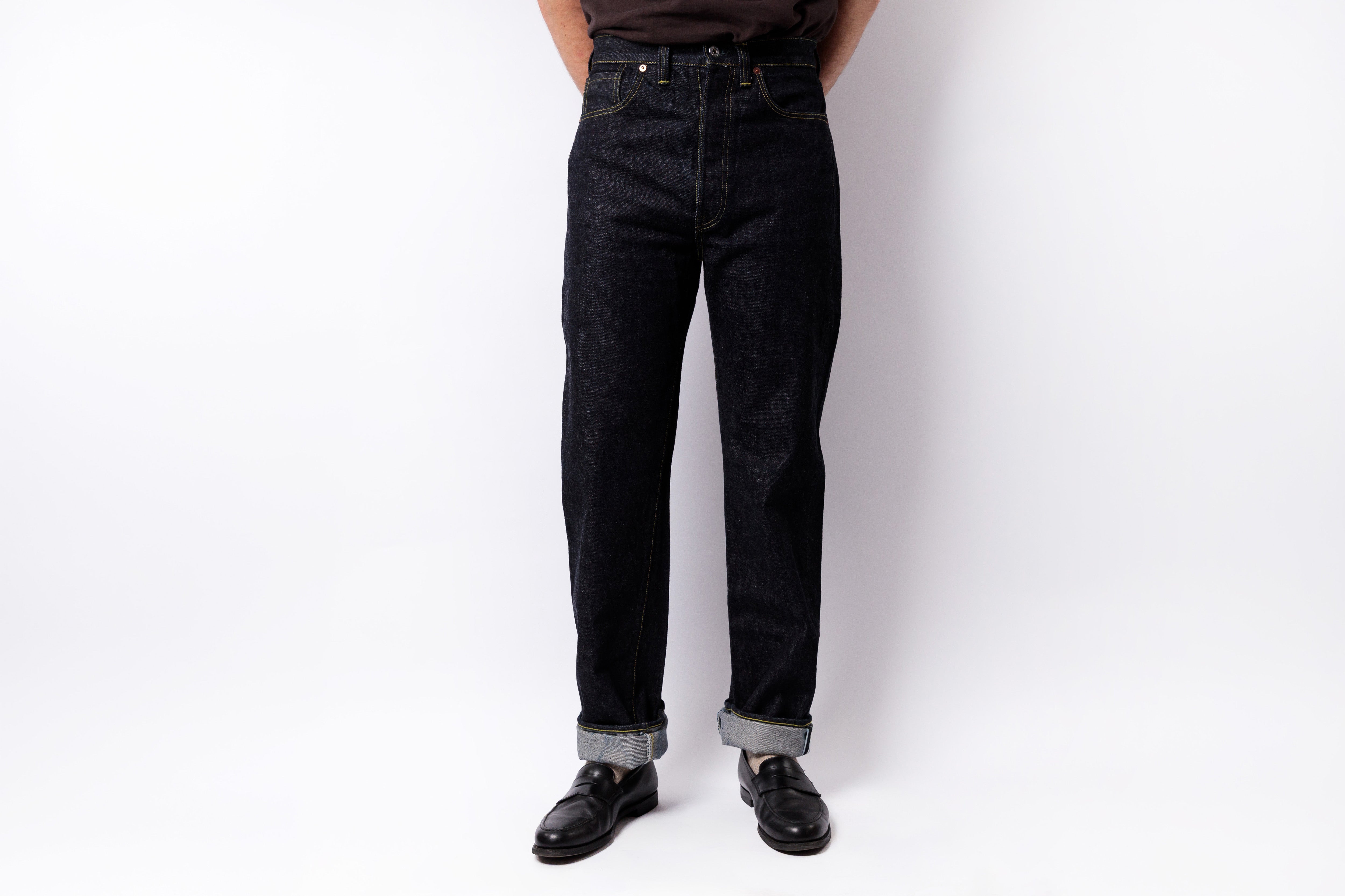 A person wearing dark indigo denim jeans with rolled cuffs, paired with black leather shoes, standing against a plain white studio background.