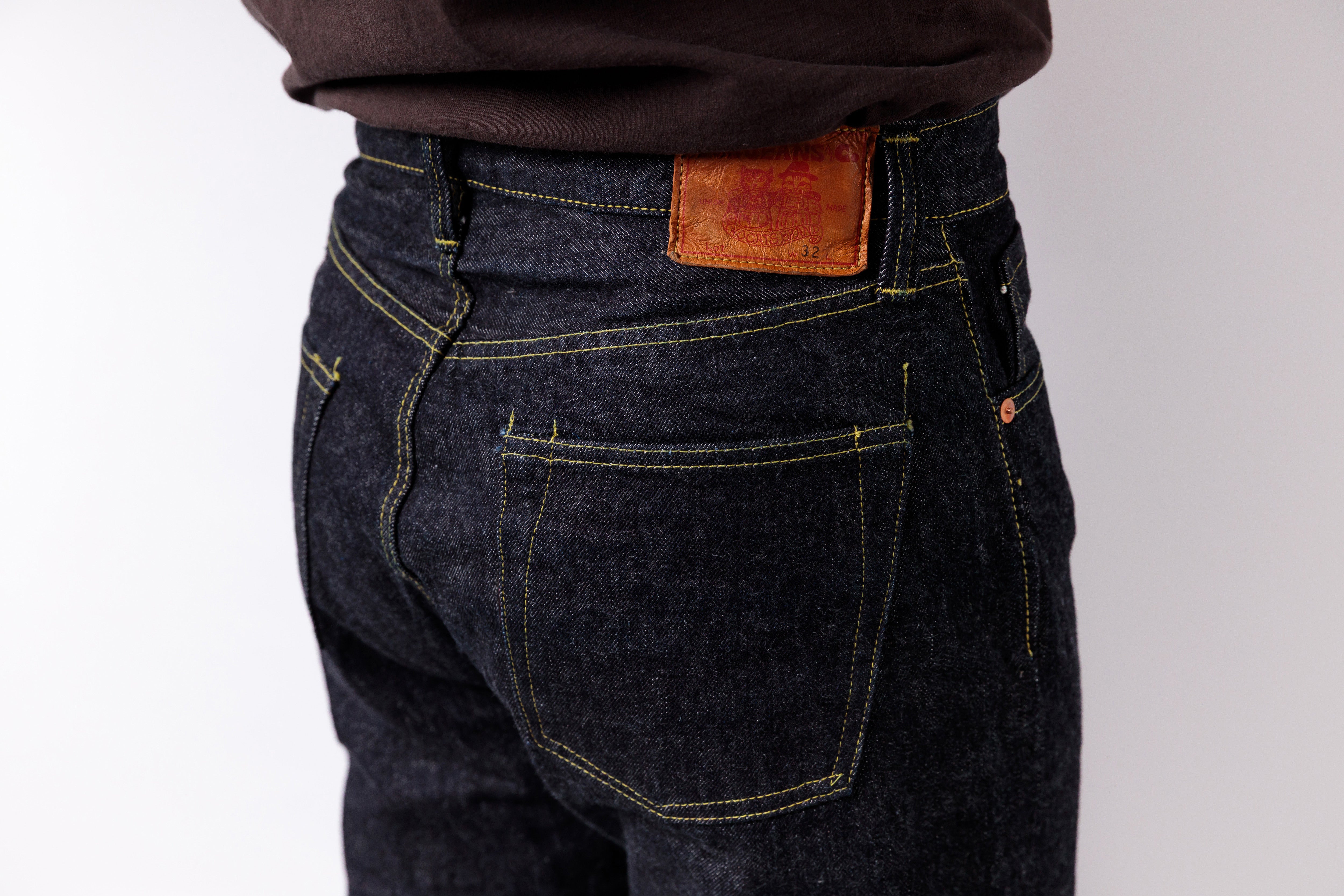 Close-up back view of a person wearing dark indigo denim jeans with yellow contrast stitching and a brown leather patch at the waistband, paired with a dark brown top against a light gray background.