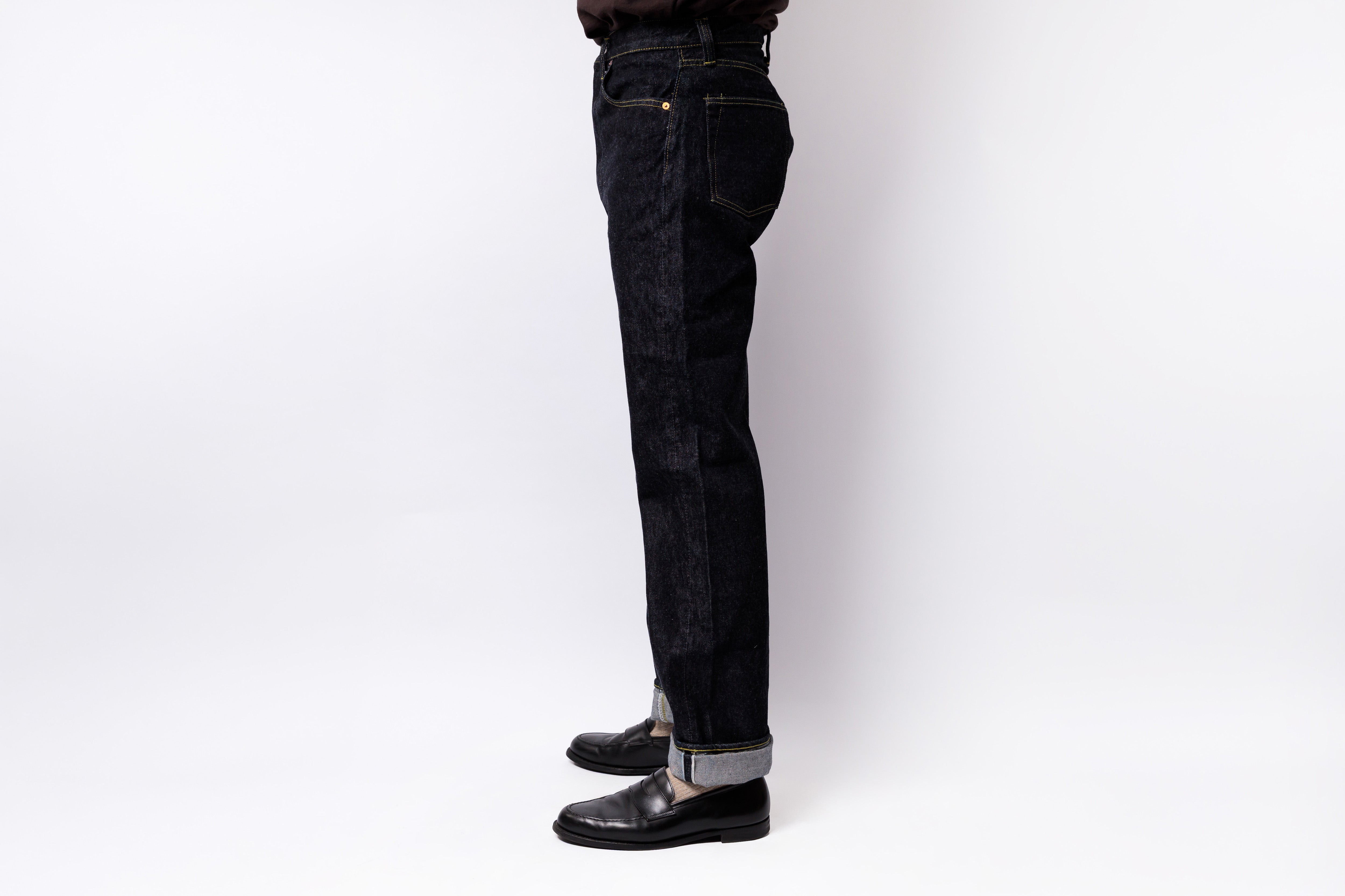 A side profile view of a person wearing dark indigo denim jeans with rolled cuffs and black leather shoes, standing against a plain white studio background.