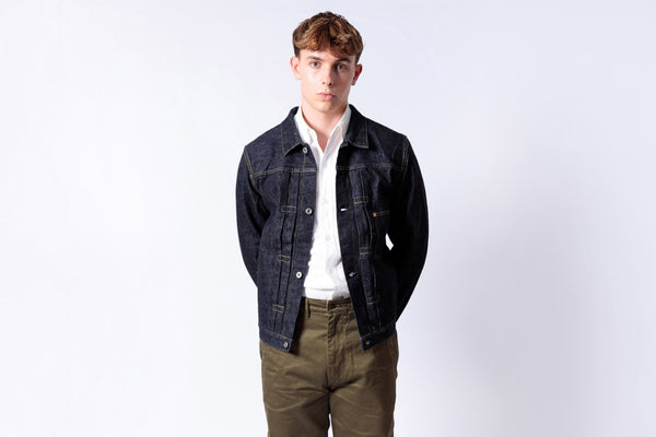 Man wearing a dark denim jacket over a white shirt and green pants on a light gray background