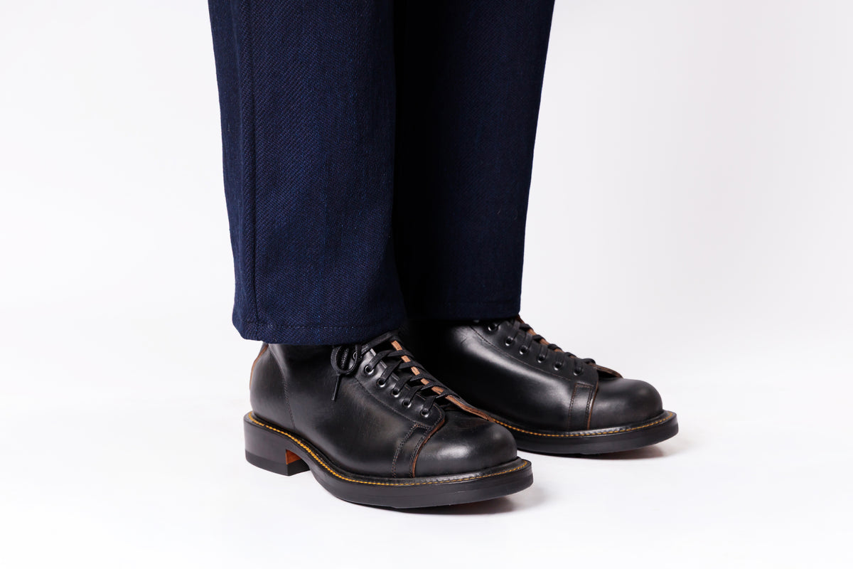 Black leather lace-up boots with a low stacked heel and contrast stitching, shown on a model wearing dark navy trousers against a white studio background.