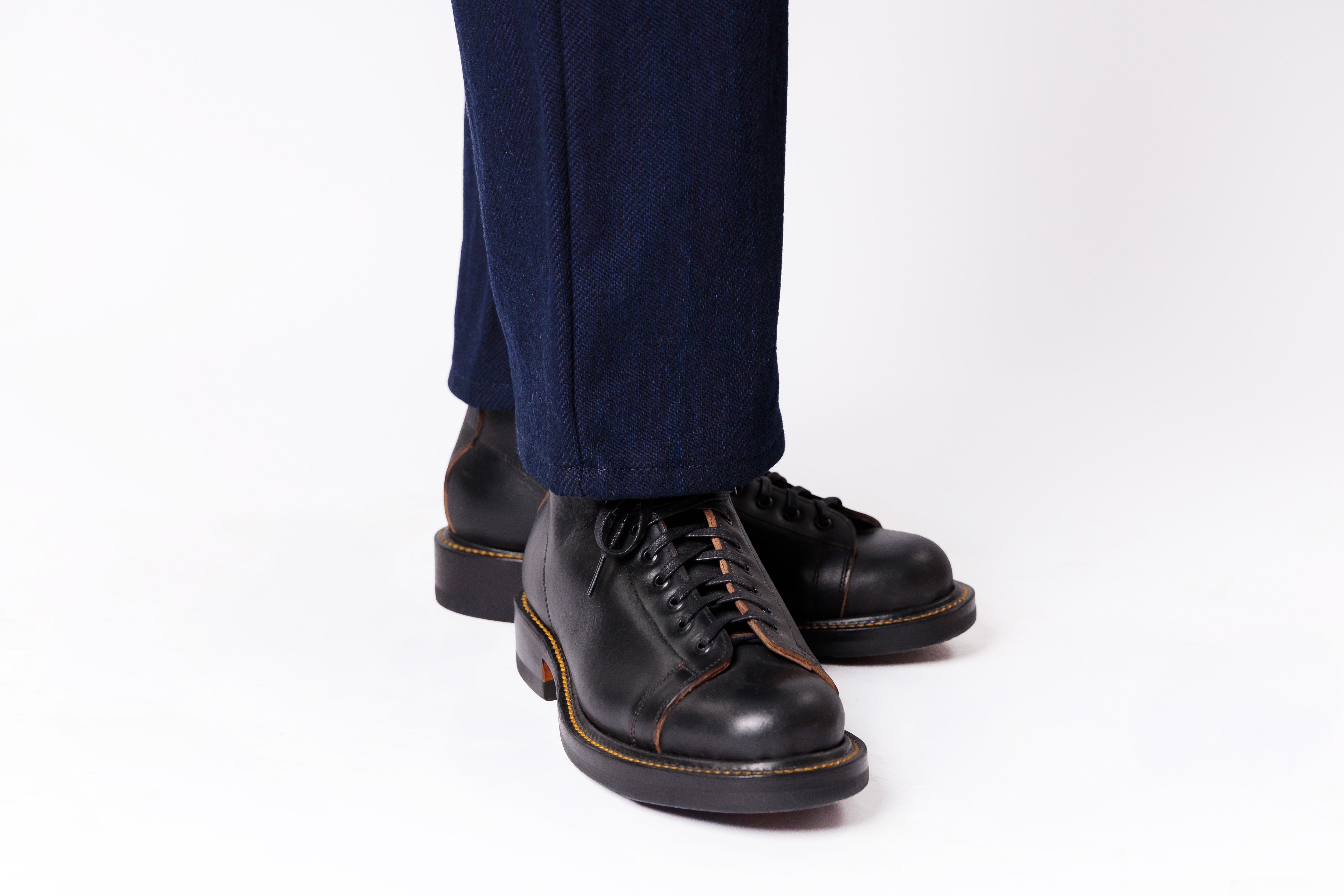 Black leather lace-up boots with contrast stitching, shown on a person's feet wearing dark blue jeans against a white studio background.