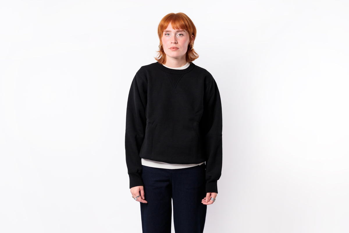 A model with short red hair wearing a black crewneck sweatshirt, a white undershirt, and dark pants, standing against a plain white background.