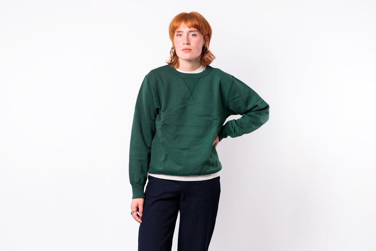 A person with short red hair wearing a dark green crewneck sweatshirt and dark navy pants, standing against a plain white background.