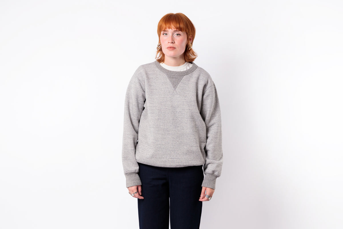 A woman with red hair wearing a light gray knit crewneck sweater with a darker gray V-neck detail, paired with dark pants, standing against a plain white background.