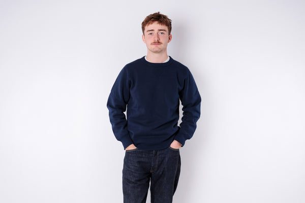 Man wearing a navy blue sweater and dark jeans against a white background